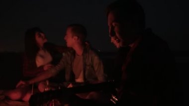 Genç insanlar şenlik ateşi ve birlikte şarkı, gitar çalmak yakınında oturuyor. Piknik, şenlik ateşi, gece