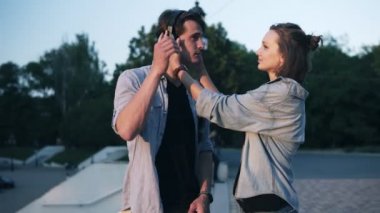 Seven genç çift akşam yumuşak güneş ışığı altında. Kız onu kulaklık erkek başını ve onu öpmek koyar. Park tarafı