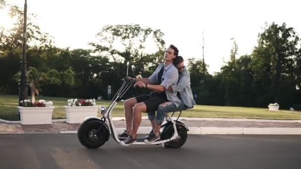 Homem confiante em camisa azul dirigindo scooter enquanto sua namorada abraçá-lo. incrível jovem mulher equitação no moped com amigo no parkside — Vídeo de Stock
