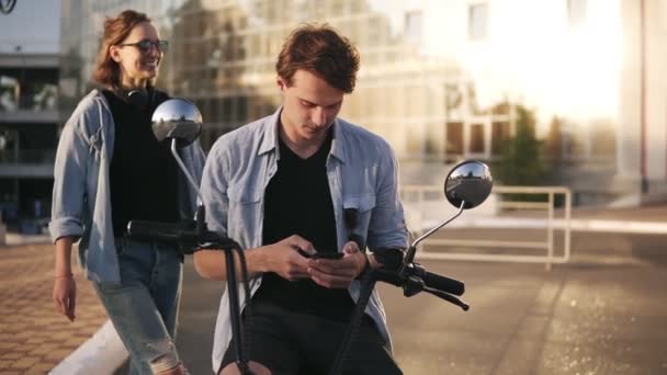 Young caucasian man is sitting on bike and scrolling his mobile phone. The attractive girlfriend coming from the back and closed his eyes with hands. Guess who. Happy, cheerful young people outdoors — Stock Video