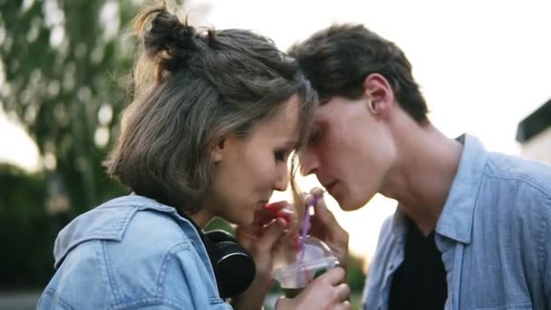 Joven pareja atractiva están bebiendo de una taza de plástico juntos. De pie en el parque y luego besándose con ternura. Movimiento lento — Vídeos de Stock
