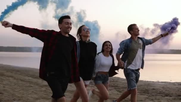 Jovens quatro pessoas correndo com granada de fumaça colorida em suas mãos junto ao mar durante o pôr do sol. Divertido, fumaça colorida em torno deles. Sorrindo, de mãos dadas — Vídeo de Stock