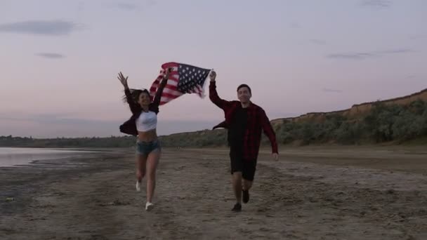 Beau jeune deux personnes courant sur un bord de mer dans la soirée. crépuscule tenant au-dessus d'un drapeau américain. Heureux, souriant, émotionnel — Video