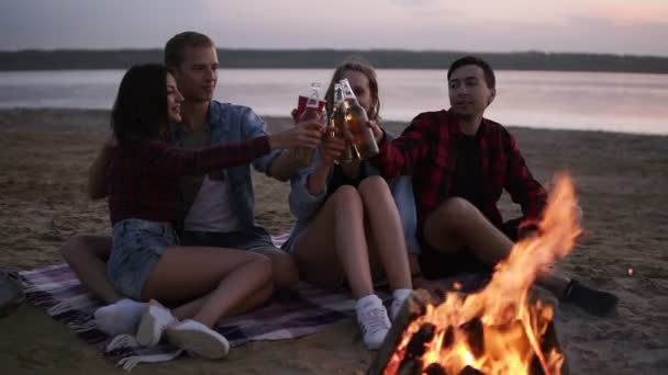 Två hipster par sitter på Pläden nära havet. Umgås tillsammans, dricka alkohol från flaskor. Skål, lycklig, leende människor nära brasan — Stockvideo