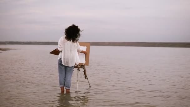 La artista femenina en jeans azules y camisa blanca de pie con caballete en el agua hasta los tobillos y trabajando en su futuro cuadro. Hermoso paisaje circundante: lago y cielo blanco claro. Vista trasera — Vídeos de Stock