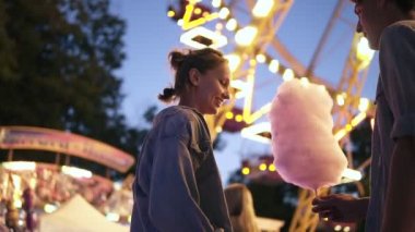 Genç çift, dating, lunaparkta toplantı erkek getirmek bir pamuk şeker kız arkadaşı. Öpüşme. Parlatıcı üzerinde arka plan parlak. Düşük açılı görüntüleri