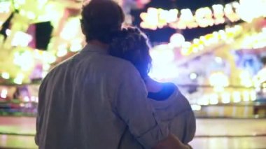 Bir eğlence parkı arcade birlikte ziyaret etti ve duran bir atlıkarınca yanındaki sarılma genç çift Geceleri ışıkları ile binmek. Arkadan ayakta