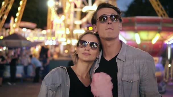 Retrato de pareja joven hipster con estilo en gafas de sol están de pie y buscando cuidadosamente las atracciones en el parque de atracciones por la noche. Comer algodón rosa de azúcar — Vídeos de Stock