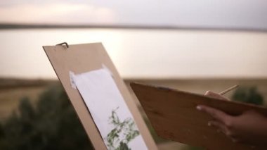 Yan görünüm açık havada bir şövale ve palet kullanarak çayır üzerinde çizim hoş bir kadın. Göl manzaralı arka planda bulanık