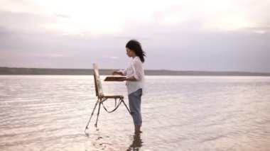 Silah, bir kız sanatçı suda ayak bilekleri çizer kadar bir şövale ve bir palet kullanarak bir manzara hız. Kıvırcık ve arka plan üzerinde kız Gölü, shes rahat kıyafetler içinde: kot pantolon ve beyaz gömlek