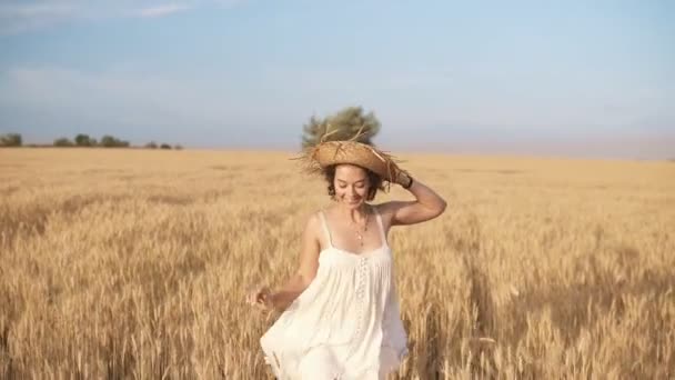 Hermosa joven en vestido blanco en el campo de trigo. Sonriente, feliz hembra corriendo y sostiene su sombrero de paja en su cabeza. Vista frontal — Vídeos de Stock