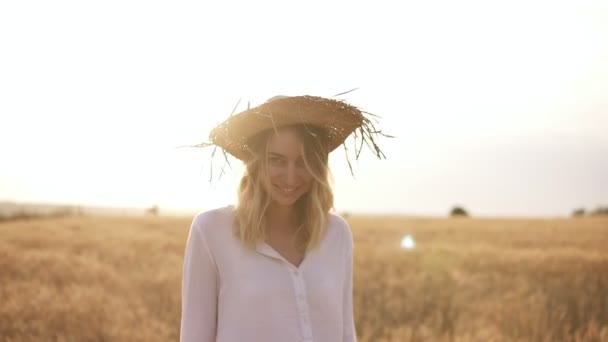 Attraktiv, rolig, leende blond kvinna i vit skjorta och stråhatt poserar medan promenader av vetefält på solig sommardag — Stockvideo