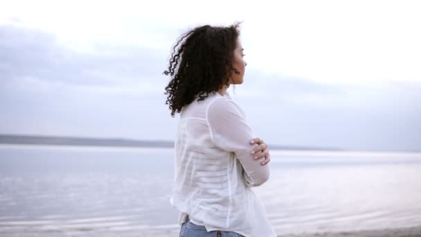 Imágenes de cerca de una atractiva joven caminando junto al mar en el agua, usando una camisa blanca. Chica morena rizada en la mañana nublada caminando al aire libre — Vídeo de stock