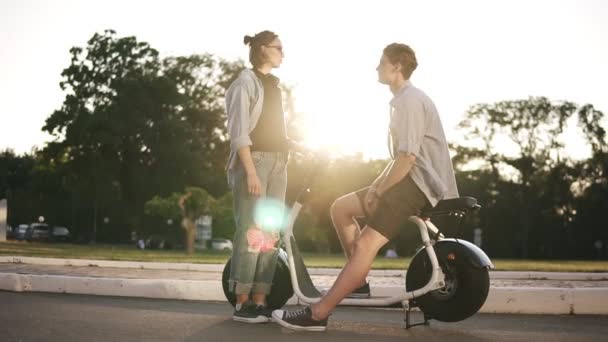 Young male and female are have time together outdoors. Boys sitting on an electro mini bike and smiling. The girl in sunglasses stands near and talks. Sun shines on the background — Stock Video