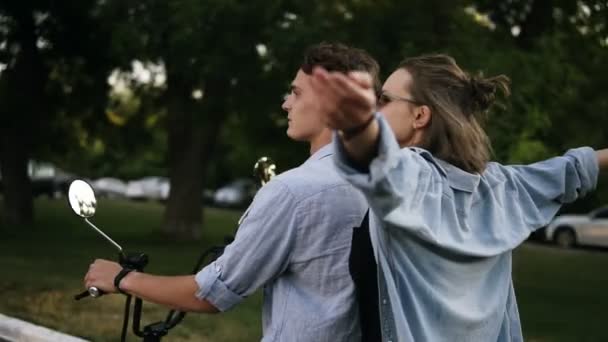 Jovem homem bonito com sua bela namorada andando de bicicleta na estrada do parque. Menina feliz, sorridente em óculos de sol, ambos em camisas azuis — Vídeo de Stock