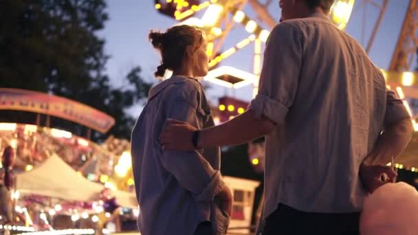 Vista de baixo ângulo de um lindo jovem casal no parque de diversões à noite. Hadsome surpreendeu sua namorada com um algodão doce. Ternura, beijando, namoro — Vídeo de Stock