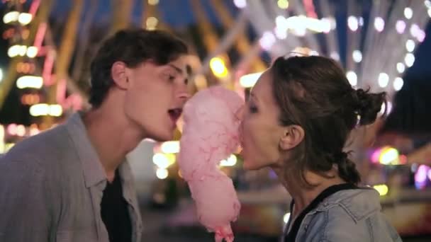 Casal jovem encantador no parque de diversões. Passar tempo juntos, comer algodão rosa doce de lados. Casal amoroso, namoro. Vista desfocada da feira de diversão em segundo plano — Vídeo de Stock