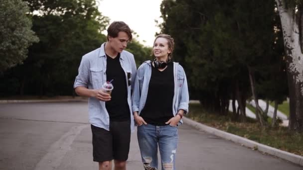 De beaux jeunes amis en t-shirts noirs et en chemises bleues se promènent dans le parc au crépuscule. Parler, sortir ensemble, l'heure d'été. Vue de face — Video