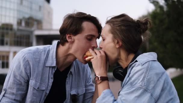 Όμορφο νεαρό ζευγάρι απολαμβάνει ένα γεύμα μαζί. Τρώει χάμπουργκερ μαζί από τις πλευρές. Βράδυ σούρουπο, πλευρά πάρκου — Αρχείο Βίντεο