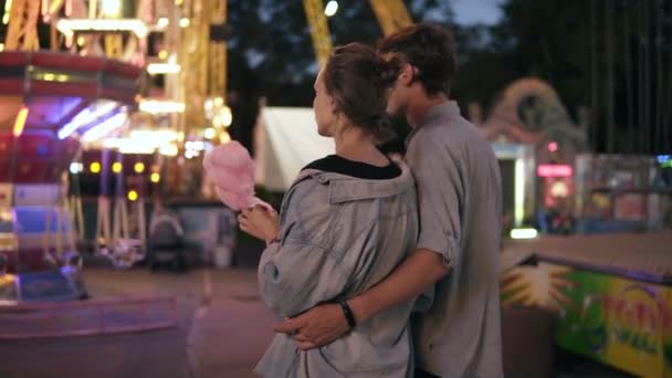 Visão traseira de um jovem namoro casal andando por diversão à noite, comer algodão doce. Ambos em camisas azuis semelhantes. Parque de diversões, par amoroso, abraço — Vídeo de Stock