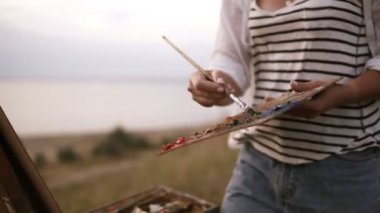 Çizgili gömlekli kız sanatçı paletteki renk karışımları ve tuval şövale üzerinde uygular. Açık havada, esen rüzgar