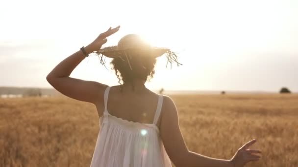 Felice, ragazza snella in cappello di paglia e vestito bianco estivo felicemente in esecuzione in campo di grano chiaro. Il sole splende sullo sfondo. Vista posteriore — Video Stock