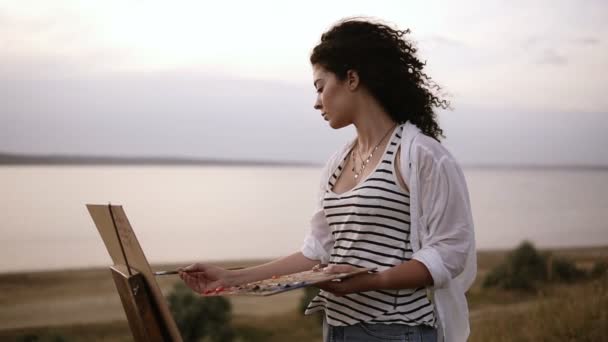 Vista lateral de uma artista mulher encaracolado atraente trabalhando em sua imagem com cavalete ao ar livre perto do lago. Manhã, vento suave soprando no rosto das mulheres — Vídeo de Stock