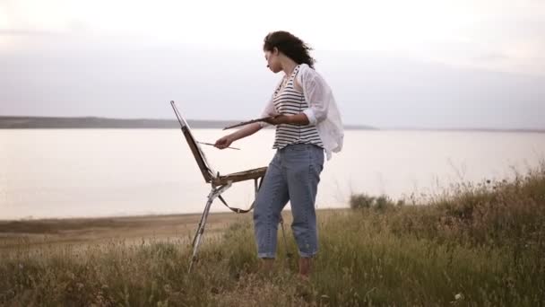 Πλήρες μήκος μήκος σε πόδηα του μια όμορφη κοπέλα με άσπρο πουκάμισο και τζιν που εργάζονται σε εξωτερικούς χώρους. Χρώματα χρησιμοποιώντας ένα καβαλέτο και λάδι χρώματα στο Λιβάδι μπροστά στη λίμνη. Άνεμος φυσά, πρωί — Αρχείο Βίντεο