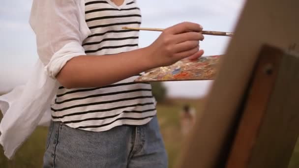 Gorgeous view of a young female artist in work process outdoors. Paints using a palette and easel. Wind waving her curly hair — Stock Video