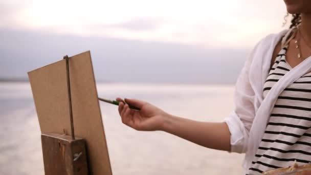 Close up of an artists art process. An easel and palette. Girl is putting some color paints on canvas. Blurred lake surround on the background