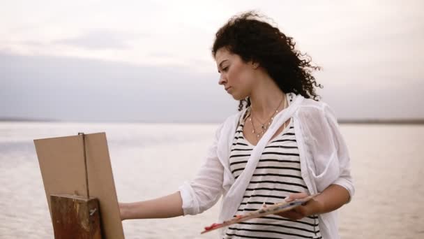 Close-up van een prachtige vrouw met brunette krullend haar is permanent aan de voorkant van het meer en tekening met behulp van een palet en een ezel. Ernstige en geconcentreerd. Ochtend heldere hemel — Stockvideo