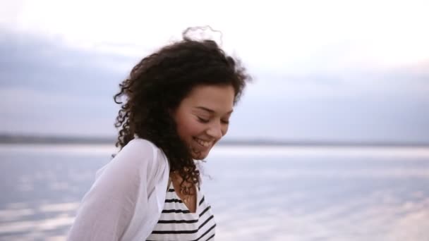 Красивая молодая кудрявая брюнетка, идущая босиком по морю или озеру. Счастливая, улыбающаяся, застенчивая женщина в полосатой футболке, белой рубашке и синих джинсах — стоковое видео