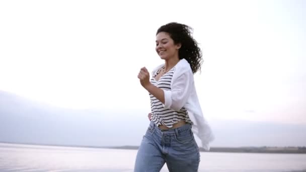 Joven, feliz, alegre chica morena felizmente corriendo por la orilla del mar en ropa casual. Libertad, estilo de vida saludable — Vídeo de stock