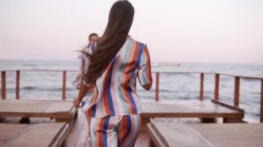 Önden görünüş muhteşem bir uzun bacaklı tabaklanmış kadınlardan tarafından ahşap sahil alanda yürüyüş. Aynı tarzda - sil renkli yaz giysileri elbise giymiş. Rüzgarlı, yaz, gündüz