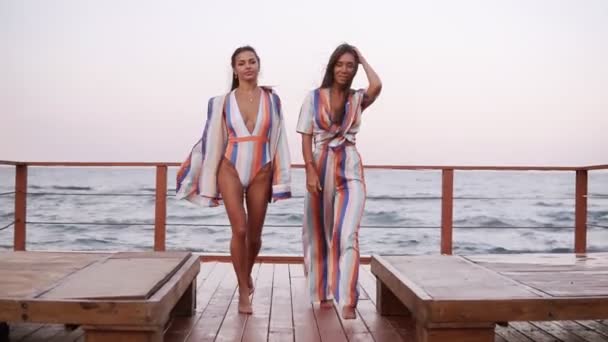 Two stylish caucasian tanned women in similar style clothes walk by wooden floor outdoors on a windy day. Seaside, front view — Stock Video