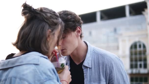 Sidovy av ungt par i blå tröjor är dring te tillsammans från en plastmugg med ett sugrör. Kvinna försiktigt kyssa en man. Utomhus — Stockvideo