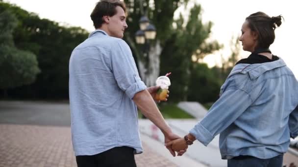 Jóvenes románticos enamorados, hombre y mujer caminando y tomados de la mano en el parque de la ciudad. Chica está bebiendo un té de una taza plásica juguetonamente. Imágenes de la parte trasera — Vídeos de Stock