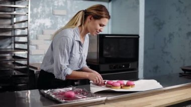 Genç gülümseyen kadın mavi gömlek ve siyah önlük hazırlama bir son sahne alanı'nda tatlı pembe camlı pasta tartles içinde koyarak - Fransız. Modern mutfak. Ekmek, pasta, mutfak konsepti