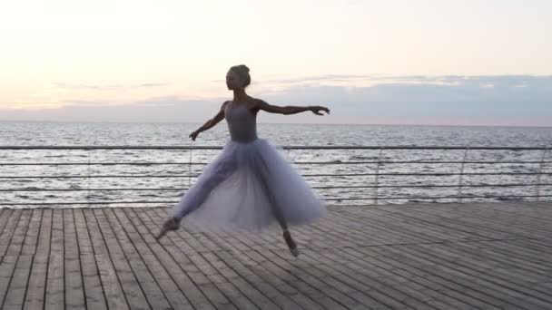 Unga vackra ballerina klädd i vit tutu dansa graciöst på sin pointe balettskor. Hoppning, utför klassiska pas på ett trägolv. Utomhus, seaside — Stockvideo