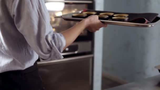 Feche as filmagens de um processo de padaria. A mulher toma uma bandeja com sobremesas e põe-na um forno prateado, moderno. Cozinha, pastelaria, conceito de sobremesas — Vídeo de Stock