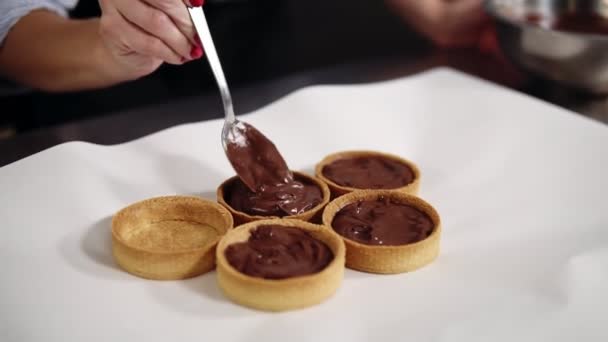 Jeune femme pâtissière préparant un dessert dans sa propre cuisine. Remplissage des tartelettes cuites au four avec de la crème au chocolat noir à l'aide d'une cuillère. Gros plan — Video