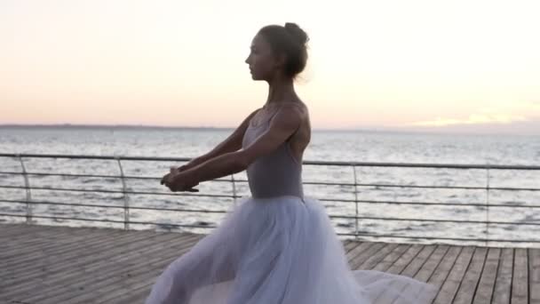 Dansende ballerina in een witte tutu en pointe op talud boven de Oceaan bij zonsopgang. Mooie jongedame beoefenen van stretching en klassieke oefeningen — Stockvideo