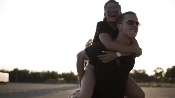 Feche as filmagens de uma cavalgada. Jovem, mulher sorridente com dreadlocks é em seus namorados de volta. Passear pela zona de estacionamento vazia. Jovens em óculos de sol se divertindo juntos — Vídeo de Stock