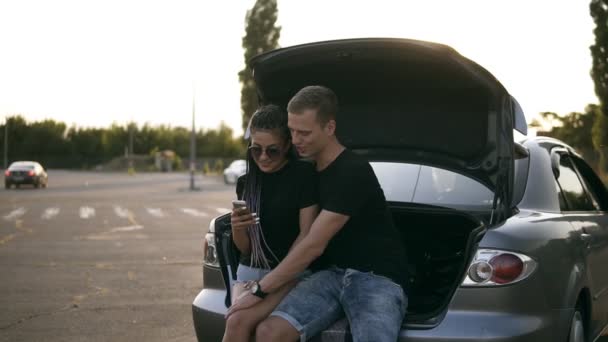 Filmagem em câmara lenta de um jovem casal caucasiano sentado no porta-bagagens de um carro aberto. Rapaz está a regatear e acariciar a namorada. Ambos procuram no smartphone. Zona de estacionamento — Vídeo de Stock