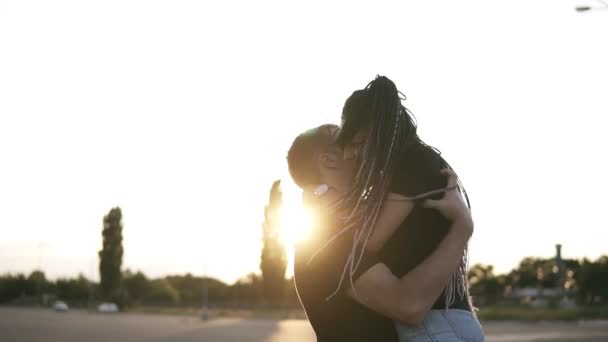 Bonito menino elegante segurando sua namorada magro em camisas jeans, de pé na estrada no entardecer verão. Bonito jovem casal abraçando e beijando em um encontro com o sol no fundo — Vídeo de Stock