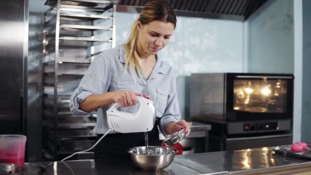 Närbild av en kvinna en konditor i en blå skjorta och en svart förkläde omrörning degen i en metall skål att lägga till ingredienser i moderna och eleganta kök — Stockvideo