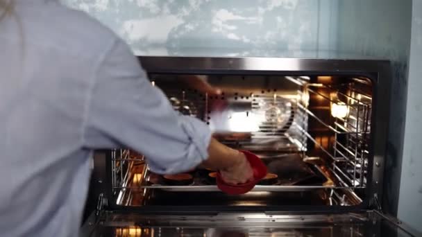 Кондитер на кухні готує приготування до десерту. Дівчина в синій сорочці тягне готові тарталетки з печі за допомогою червоної силіконової рукавички — стокове відео
