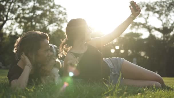 На відкритому повітрі портрет двох друзів-жінки, які приймають селфі зі смартфоном. Дві дівчини розважаються в парку з маленькою чорно-білою собакою. зелена трава і сонце світить. Повільний рух — стокове відео