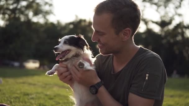 El hombre se sienta en la hierba en el parque verde, jugar con el perrito joven. Propietario burla perrito, jugar y abrazo. Sonriente, feliz propietario divirtiéndose con su mascota. Movimiento lento — Vídeos de Stock