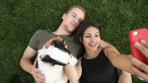 Joven, atractiva pareja caucásica están acostados en la hierba verde en el parque. El hombre sostiene a su pequeño perro blanco y negro mientras su novia morena toma fotos con su teléfono inteligente. Filmación — Vídeo de stock